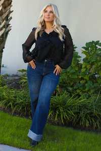 Woman model wearing a black top and blue jeans standing outdoors with greenery in the background.