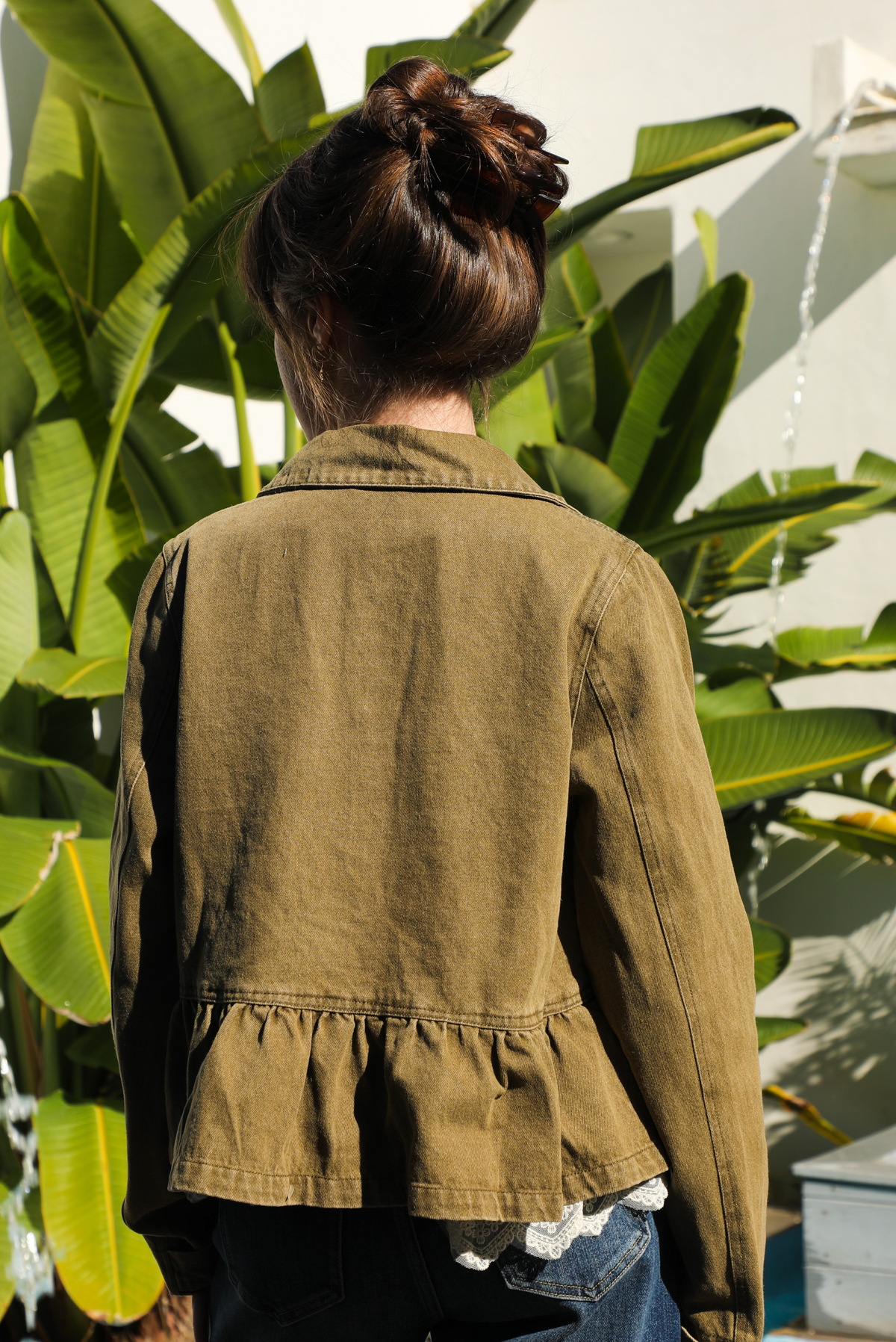 Back of a woman model outdoors wearing an olive denim jacket, cream lace top, and dark denim jeans. 