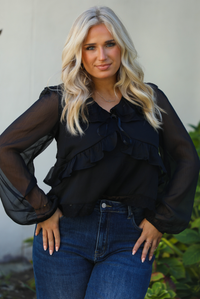 Close up of a woman model wearing a black top and blue jeans standing outdoors with greenery in the background.
