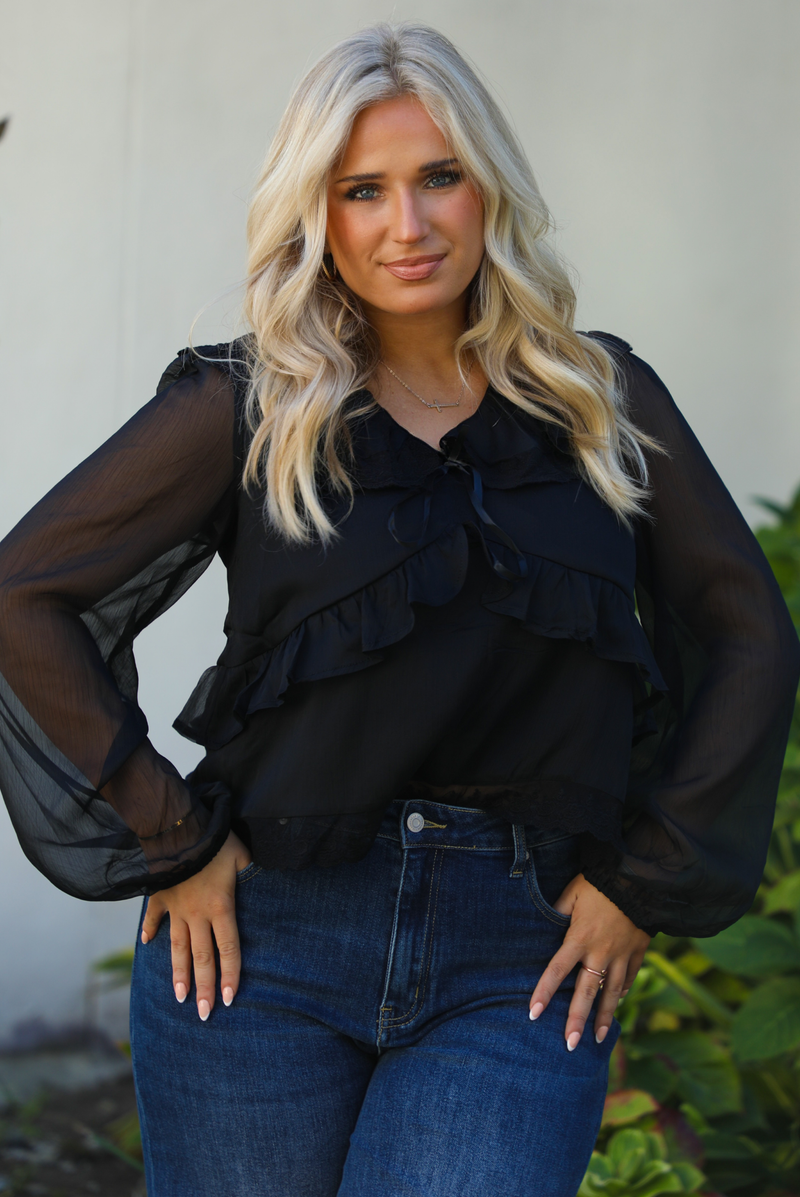 Close up of a woman model wearing a black top and blue jeans standing outdoors with greenery in the background.