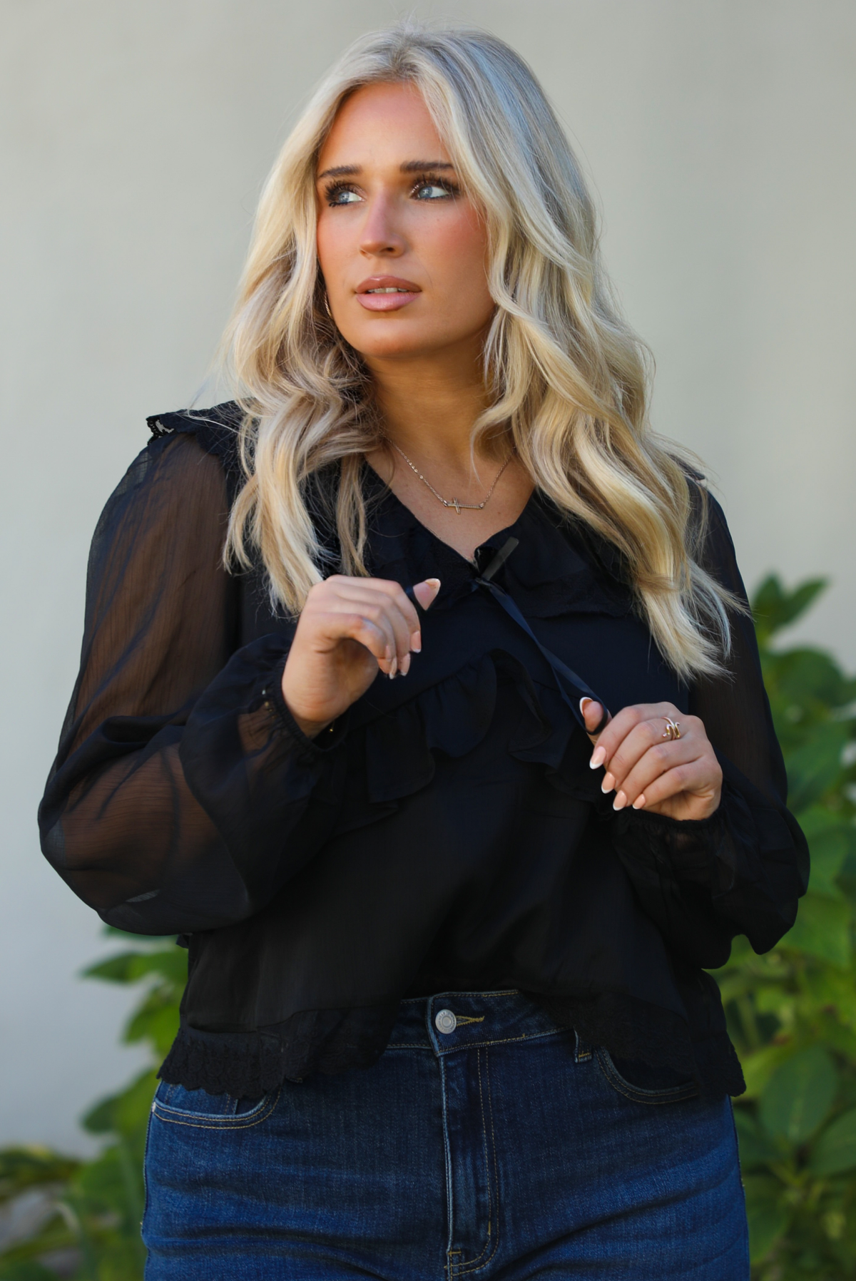 Close up of a woman model wearing a black top and blue jeans standing outdoors with greenery in the background.
