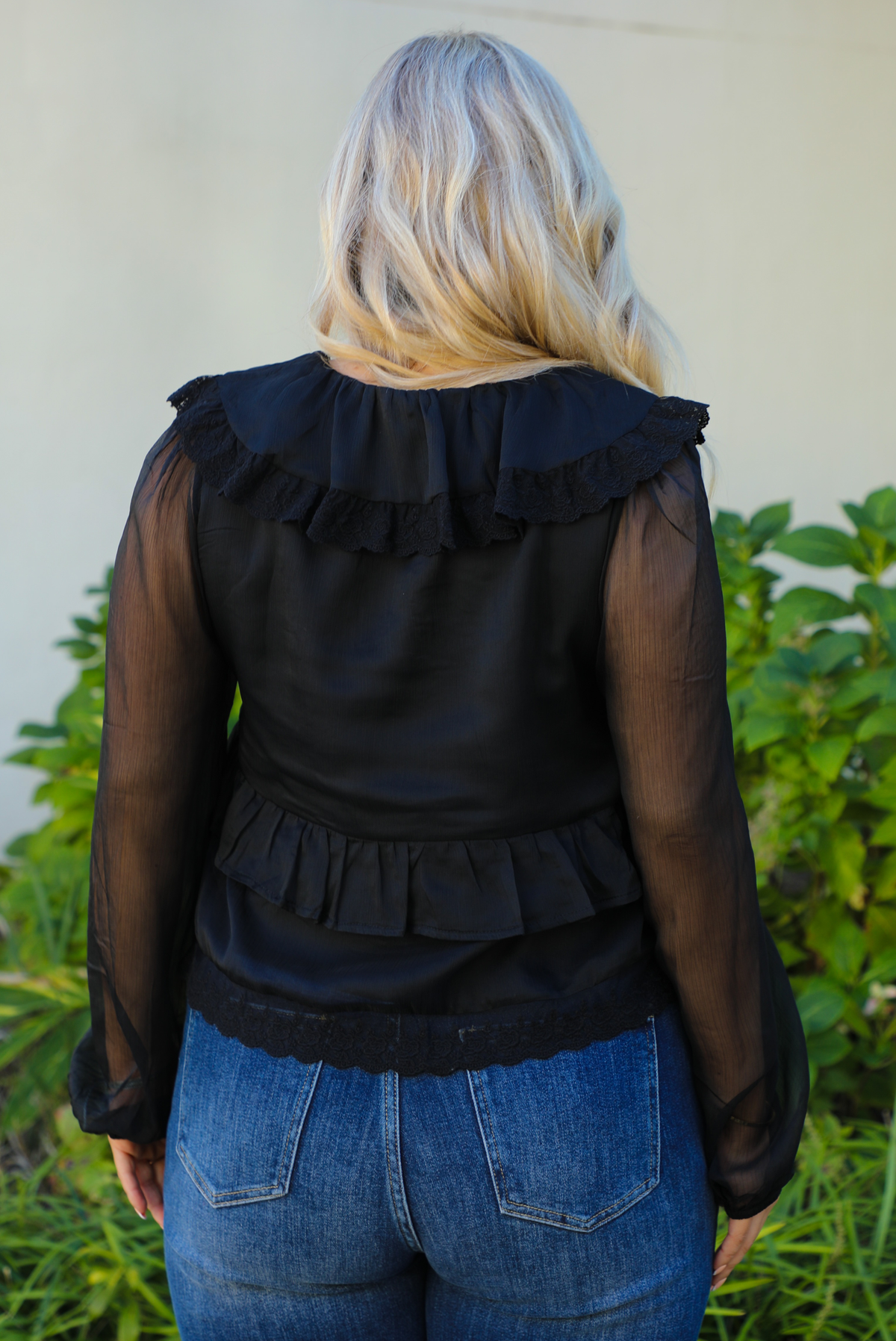 Close up of the back of a woman model wearing a black top and blue jeans standing outdoors with greenery in the background.