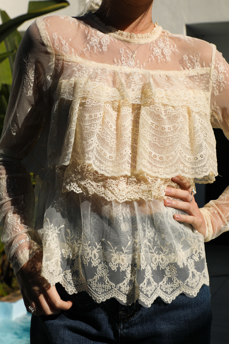 Woman model wearing a cream lace top and dark denim jeans outdoors against greenery and a water feature. 