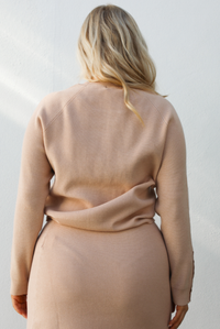 Woman model wearing a tan colored two piece set that features a long sleeve top and a midi length skirt with mustard colored boots. The photo showcases the back of the top and is taken outdoors against a white wall. 