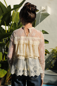 Woman model wearing a cream lace top and dark denim jeans outdoors against greenery and a water feature. 