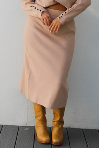 Woman model wearing a tan colored two piece set that features a long sleeve top and a midi length skirt with mustard colored boots. The photo showcases the skirt a bit better and is taken outdoors against a white wall. 