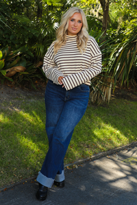 Woman model outdoors wearing a cream and burgundy striped sweater and dark denim jeans with greenery in the back. 