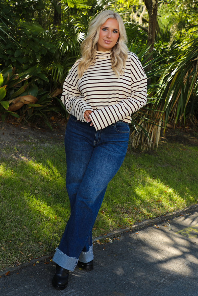Woman model outdoors wearing a cream and burgundy striped sweater and dark denim jeans with greenery in the back. 