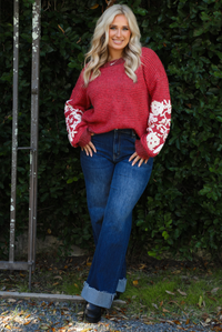 Woman model wearing a red and burgundy striped sweater with a cream colored embroidered floral pattern on the sleeves. The sweater is paired with dark denim jeans. The photo is taken outdoors against greenery. 
