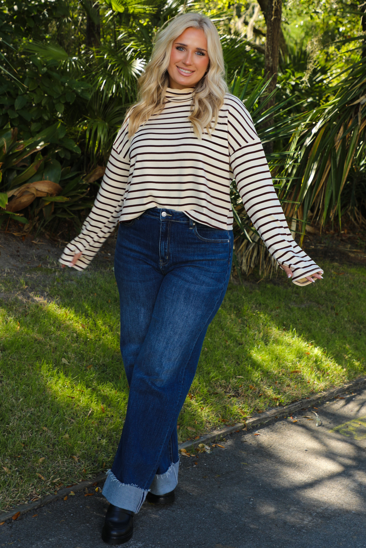 Woman model outdoors wearing a cream and burgundy striped sweater and dark denim jeans with greenery in the back. 