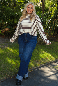 Woman model outdoors wearing a cream and burgundy striped sweater and dark denim jeans with greenery in the back. 