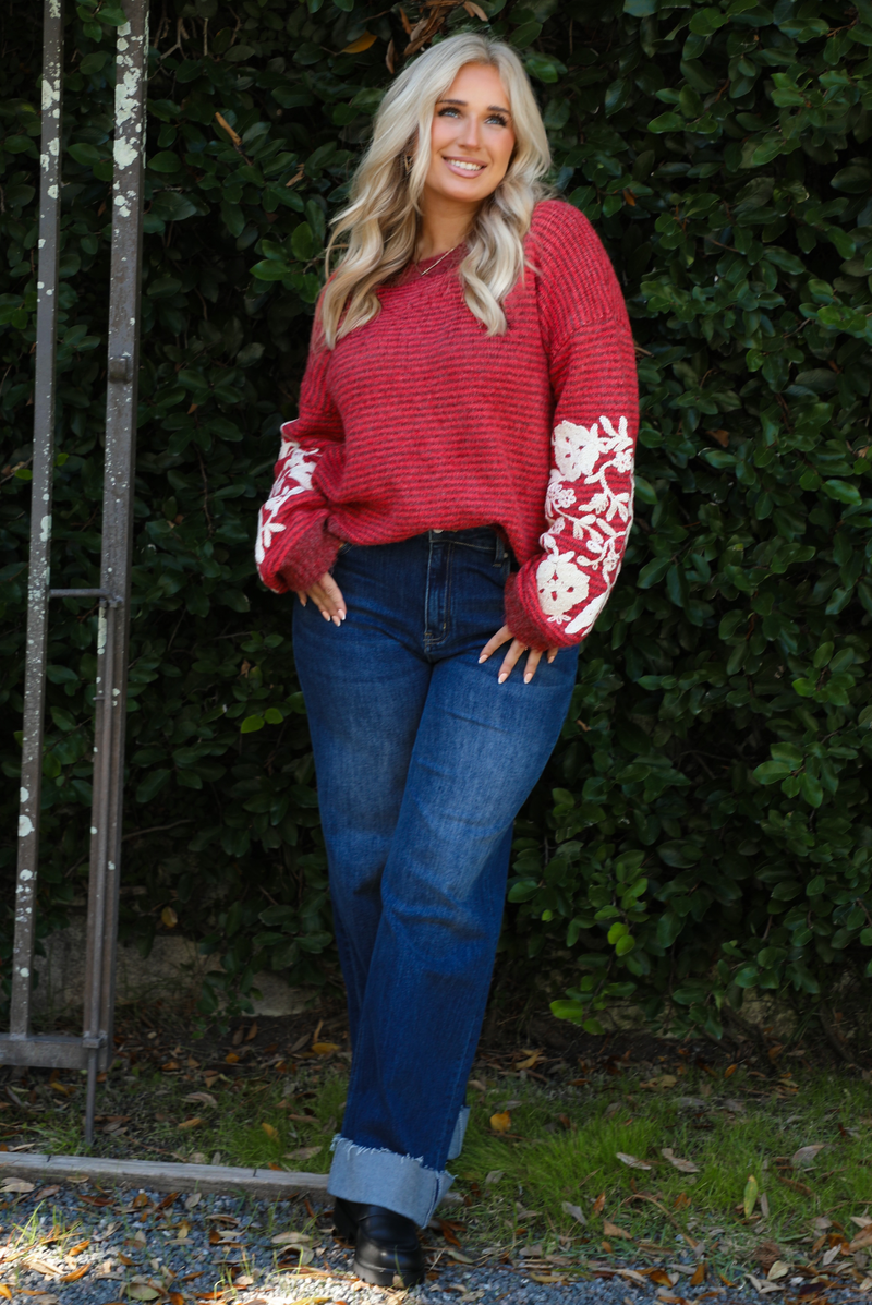 Woman model wearing a red and burgundy striped sweater with a cream colored embroidered floral pattern on the sleeves. The sweater is paired with dark denim jeans. The photo is taken outdoors against greenery. 