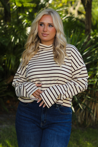 Woman model outdoors wearing a cream and burgundy striped sweater and dark denim jeans with greenery in the back. 