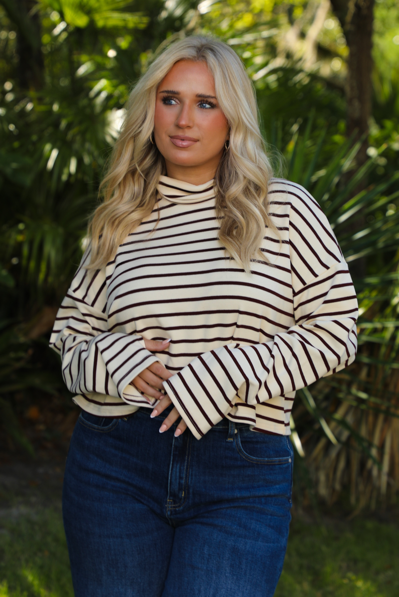 Woman model outdoors wearing a cream and burgundy striped sweater and dark denim jeans with greenery in the back. 