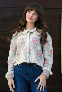Close up of a woman model outdoors against a dark brown door wearing a cream and multi-colored floral print cardigan and dark denim jeans. The cardigan has a collared neckline, functional button closures in the front, and a crimped hem. 