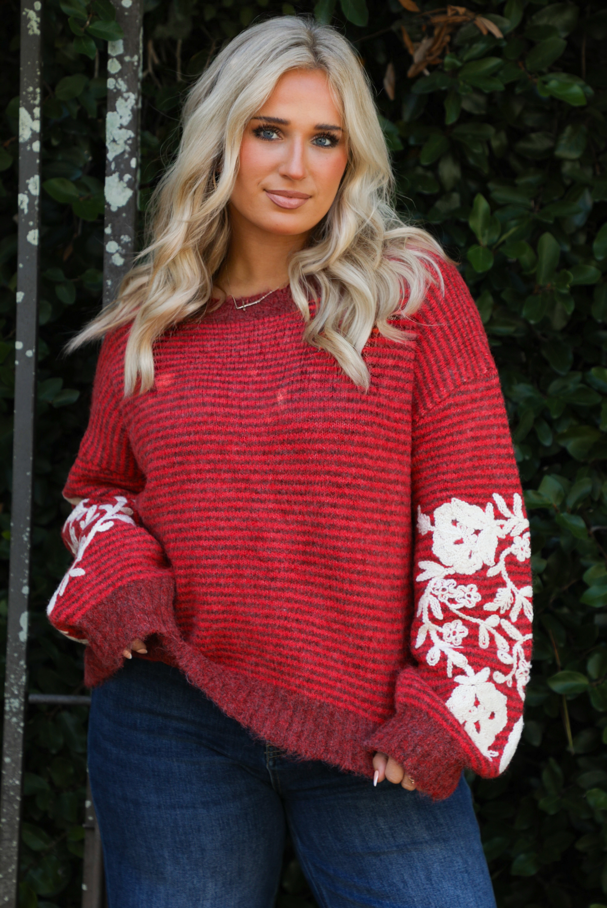 Close up of a woman model wearing a red and burgundy striped sweater with a cream colored embroidered floral pattern on the sleeves. The sweater is paired with dark denim jeans. The photo is taken outdoors against greenery. 