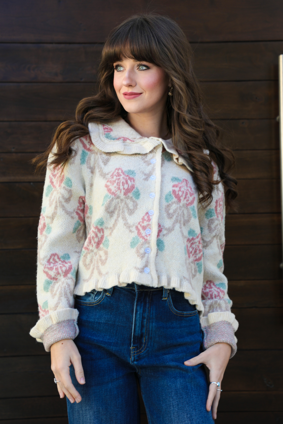 Close up of a woman model outdoors against a dark brown door wearing a cream and multi-colored floral print cardigan and dark denim jeans. The cardigan has a collared neckline, functional button closures in the front, and a crimped hem. 
