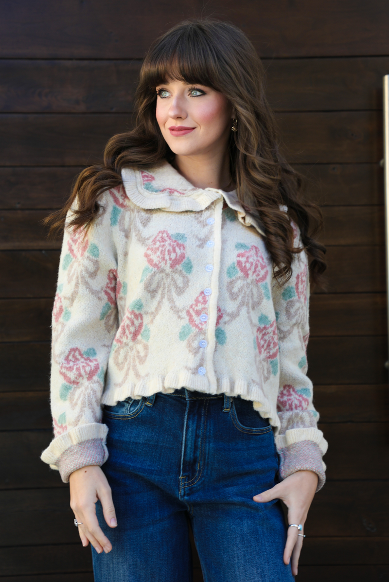 Close up of a woman model outdoors against a dark brown door wearing a cream and multi-colored floral print cardigan and dark denim jeans. The cardigan has a collared neckline, functional button closures in the front, and a crimped hem. 