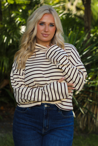 Woman model outdoors wearing a cream and burgundy striped sweater and dark denim jeans with greenery in the back. 