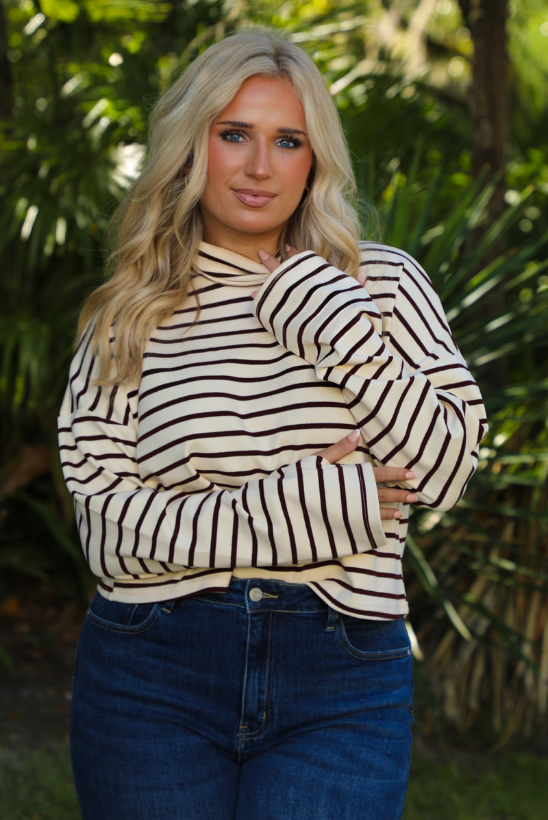 Woman model outdoors wearing a cream and burgundy striped sweater and dark denim jeans with greenery in the back. 