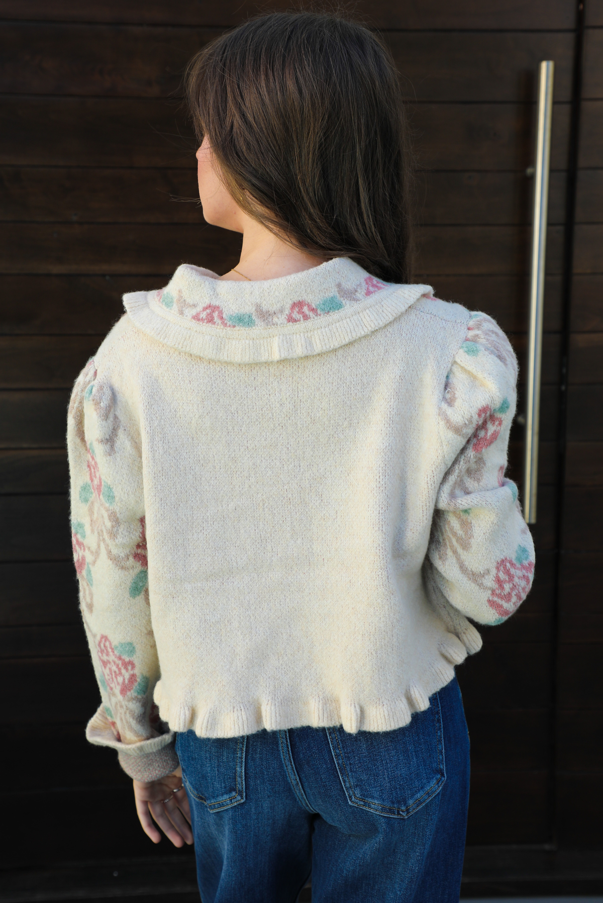 Close up of the back of a woman model outdoors against a dark brown door wearing a cream and multi-colored floral print cardigan and dark denim jeans. The cardigan has a collared neckline, functional button closures in the front, and a crimped hem. 