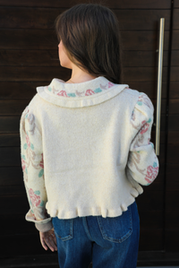 Close up of the back of a woman model outdoors against a dark brown door wearing a cream and multi-colored floral print cardigan and dark denim jeans. The cardigan has a collared neckline, functional button closures in the front, and a crimped hem. 
