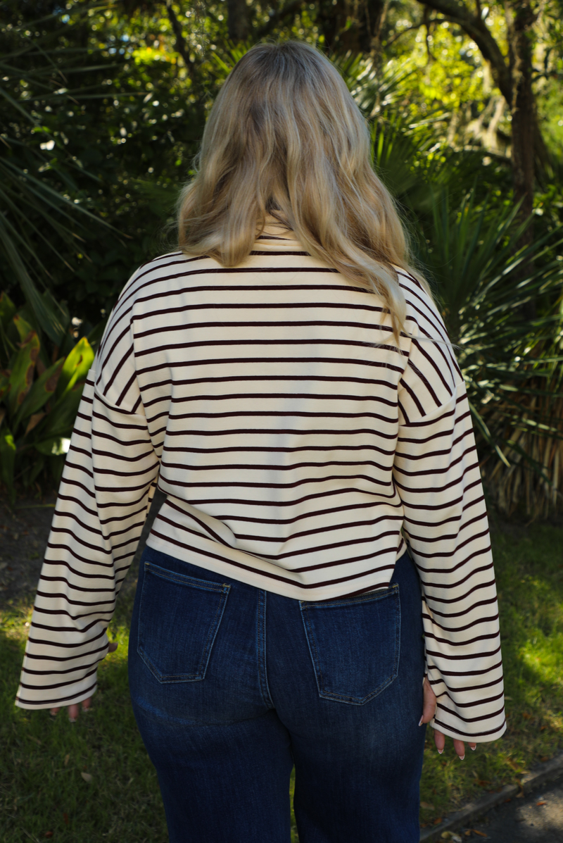 Back of a woman model outdoors wearing a cream and burgundy striped sweater and dark denim jeans with greenery in the back. 