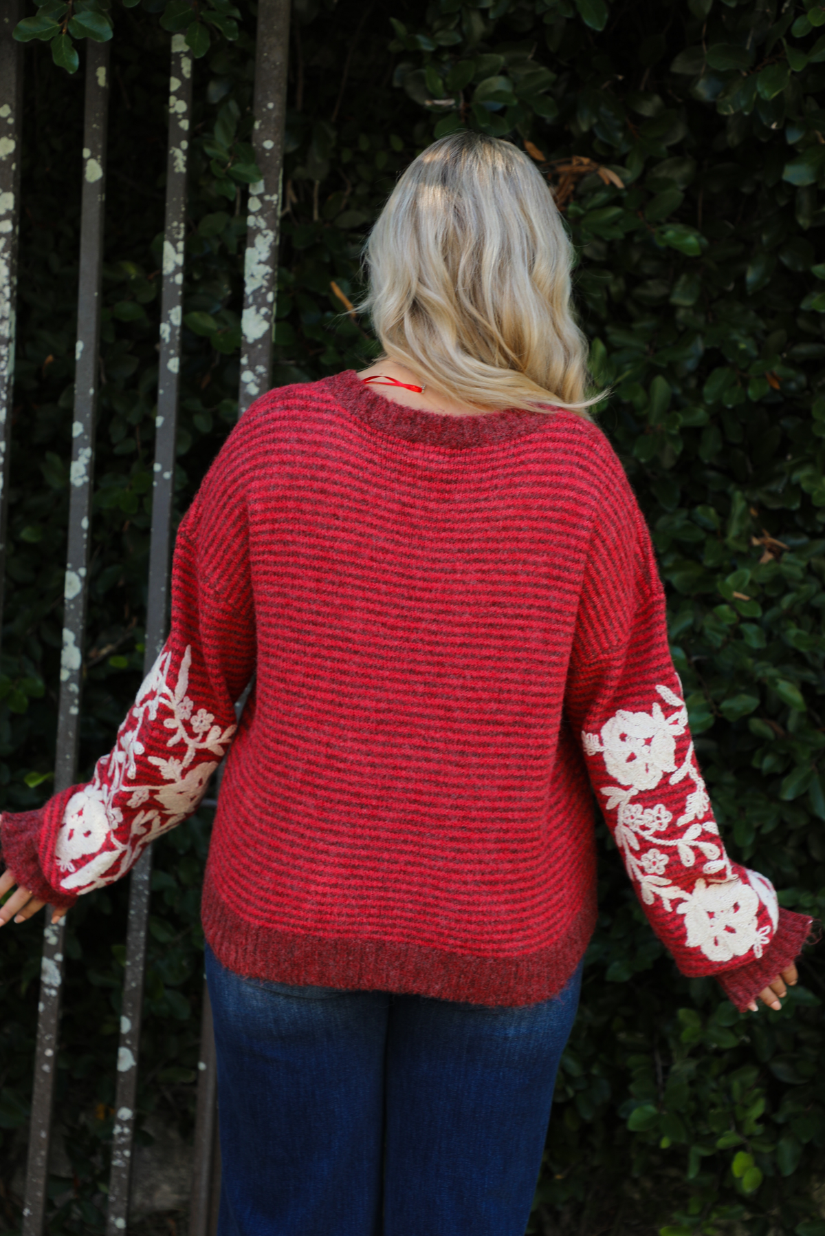 Close up of the back of a woman model wearing a red and burgundy striped sweater with a cream colored embroidered floral pattern on the sleeves. The sweater is paired with dark denim jeans. The photo is taken outdoors against greenery. 
