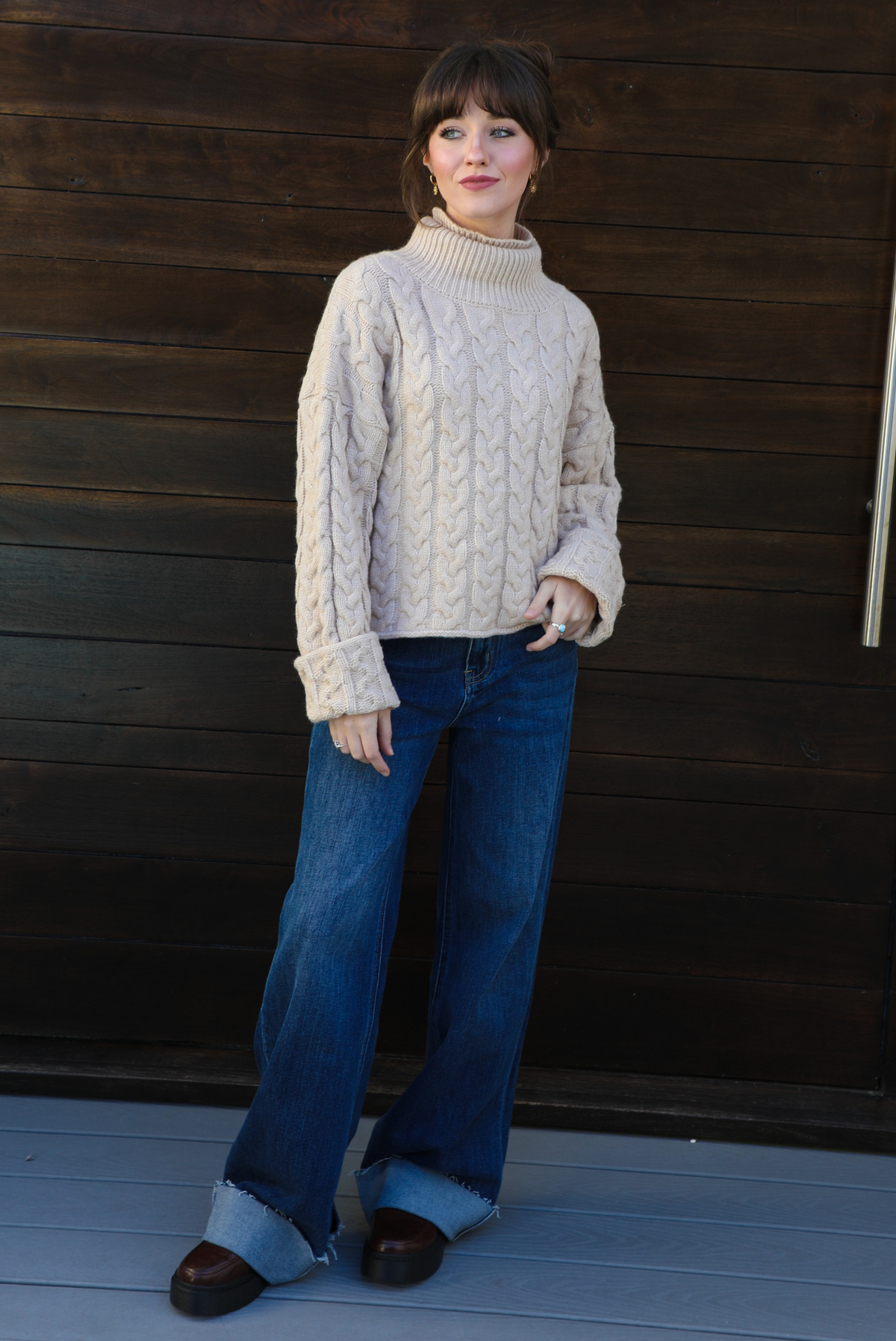 Woman model wearing a tan, thick, cable knit turtle neck sweater with dark denim jeans outdoors in front of a dark stained wooden door. 