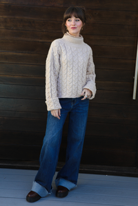 Woman model wearing a tan, thick, cable knit turtle neck sweater with dark denim jeans outdoors in front of a dark stained wooden door. 
