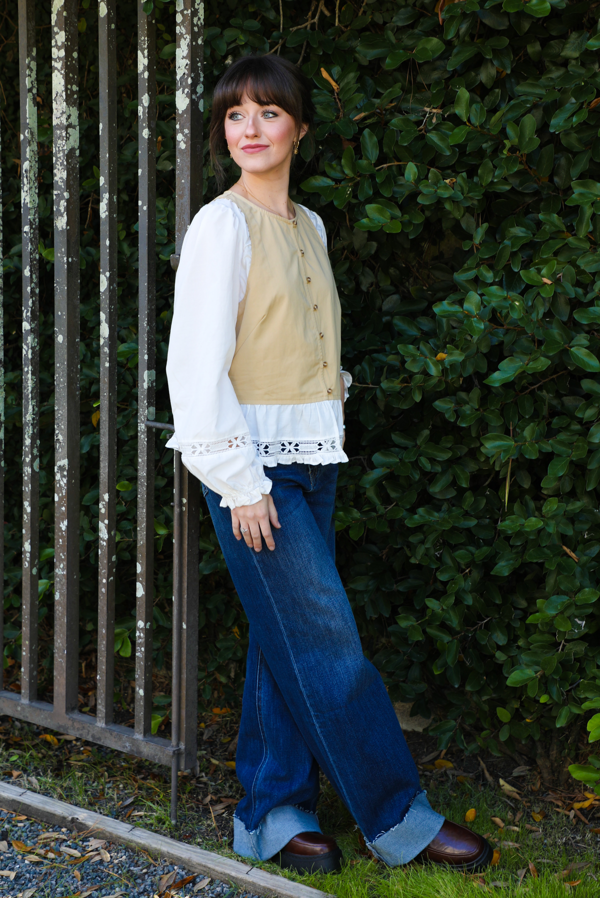Woman model wearing a tan and white blouse with lace details and blue jeans standing against a green hedge.