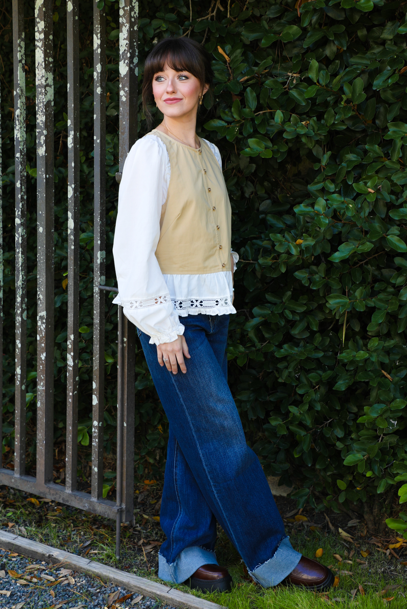 Woman model wearing a tan and white blouse with lace details and blue jeans standing against a green hedge.