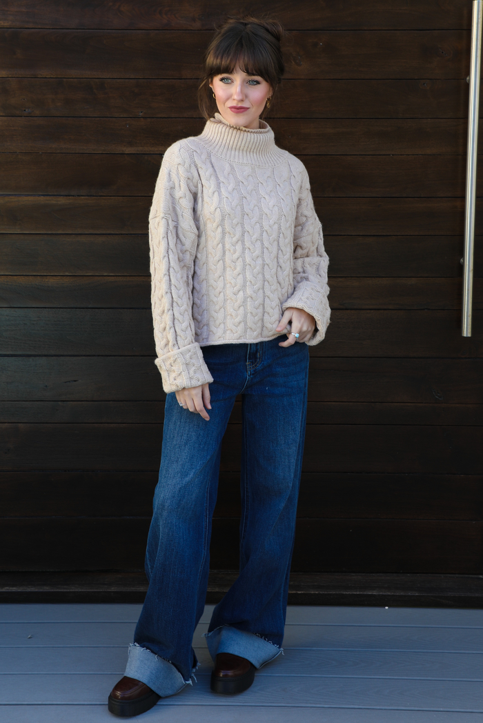 Woman model wearing a tan, thick, cable knit turtle neck sweater with dark denim jeans outdoors in front of a dark stained wooden door. 