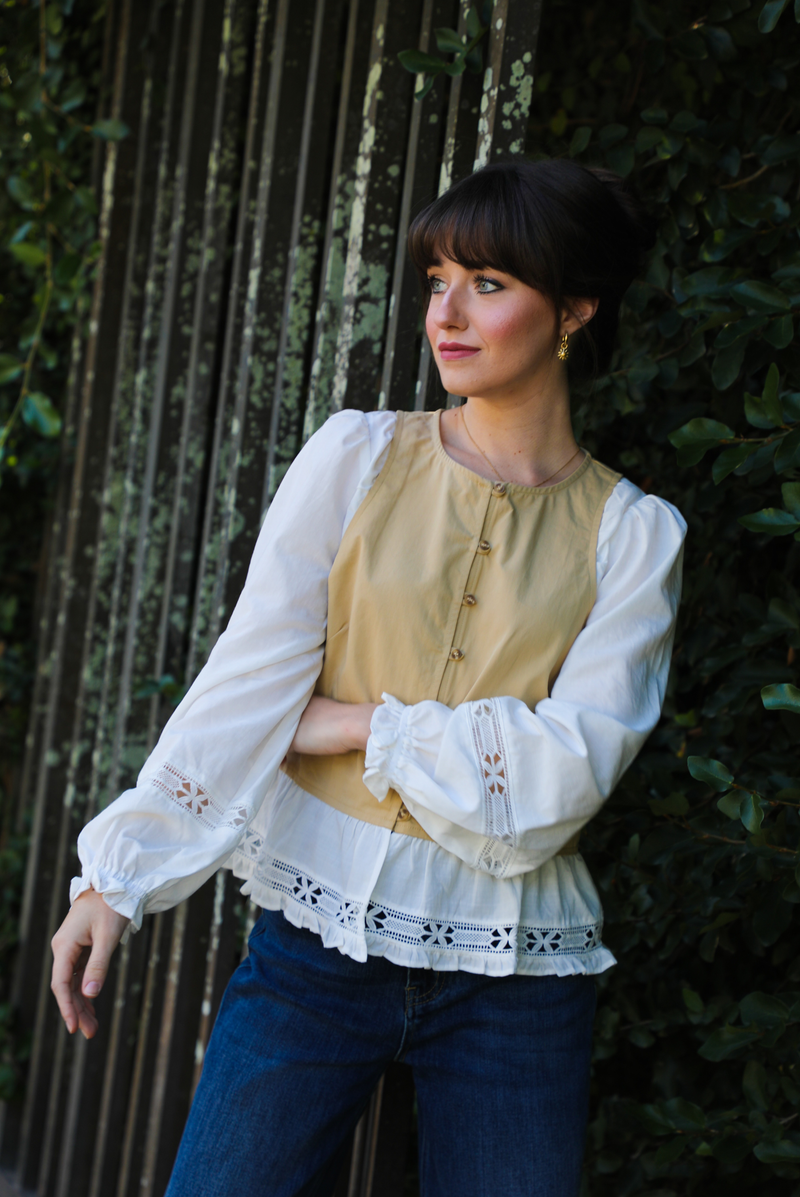 Woman model wearing a tan and white blouse with lace details and blue jeans standing against a green hedge.