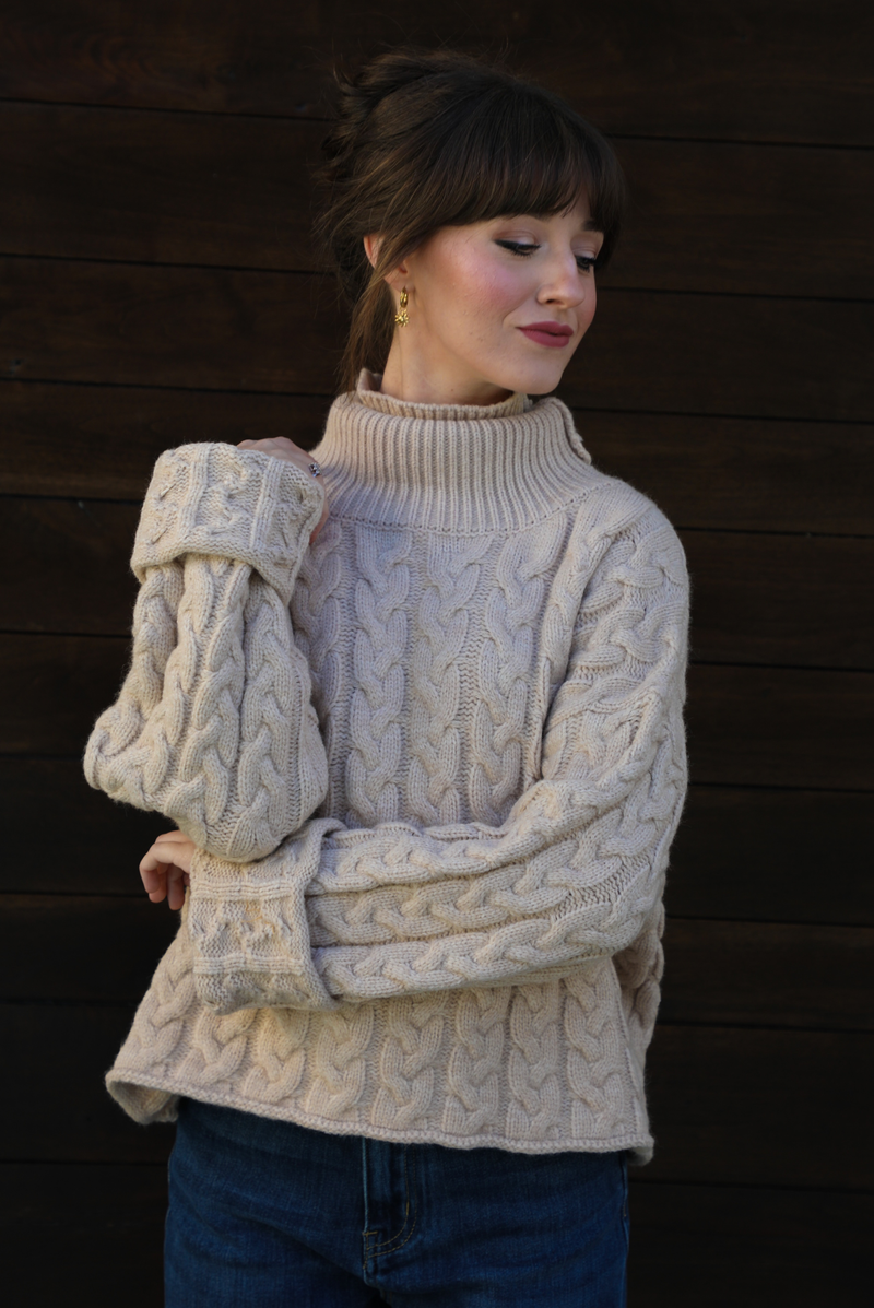 Woman model wearing a tan, thick, cable knit turtle neck sweater with dark denim jeans outdoors in front of a dark stained wooden door. 