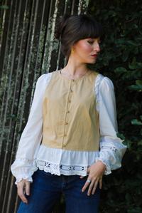 Woman model wearing a tan and white blouse with lace details and blue jeans standing against a green hedge.