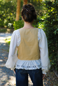 Back of a woman model wearing a tan and white blouse with lace trim details and dark denim jeans. The photo is taken outdoors with a gravel driveway and greenery around. 