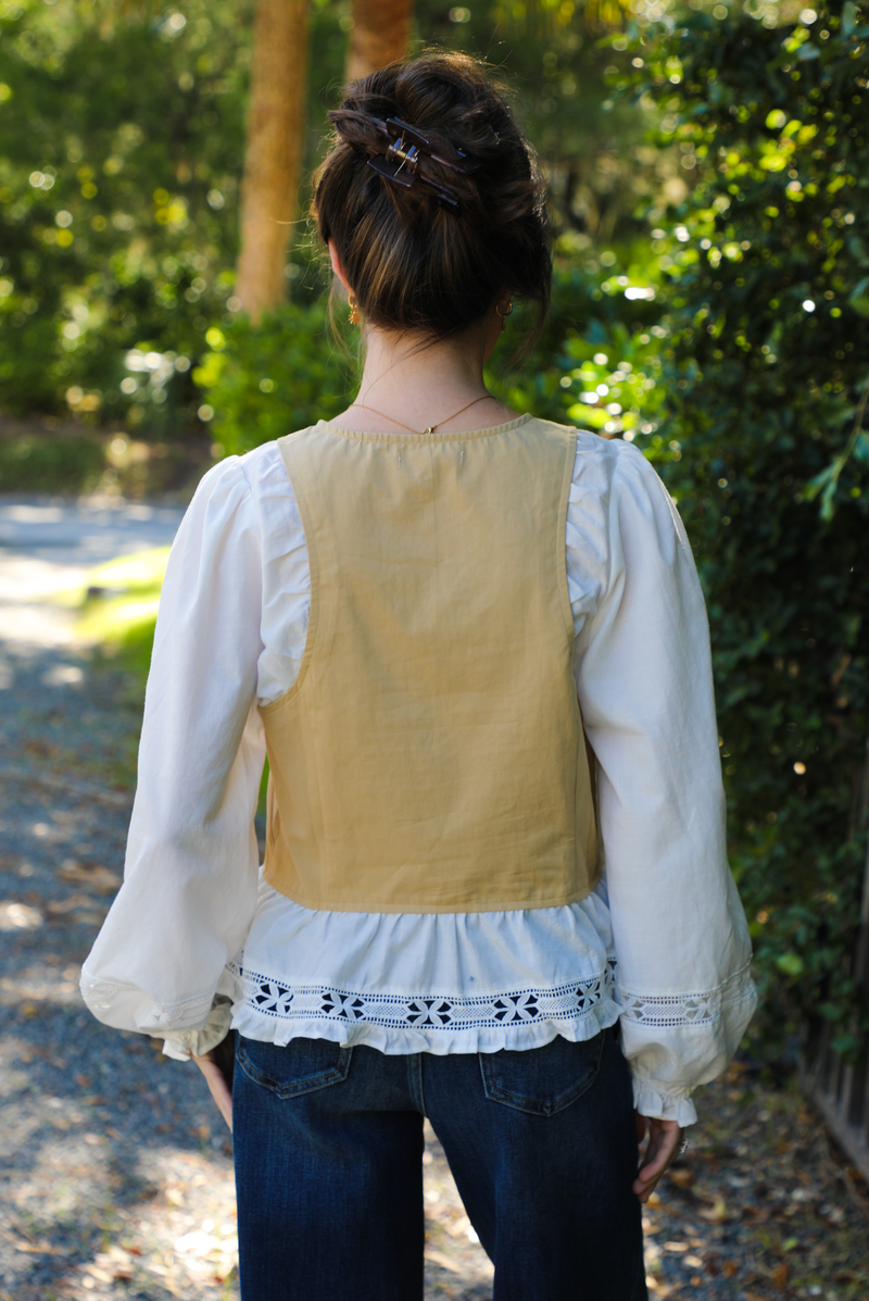 Back of a woman model wearing a tan and white blouse with lace trim details and dark denim jeans. The photo is taken outdoors with a gravel driveway and greenery around. 