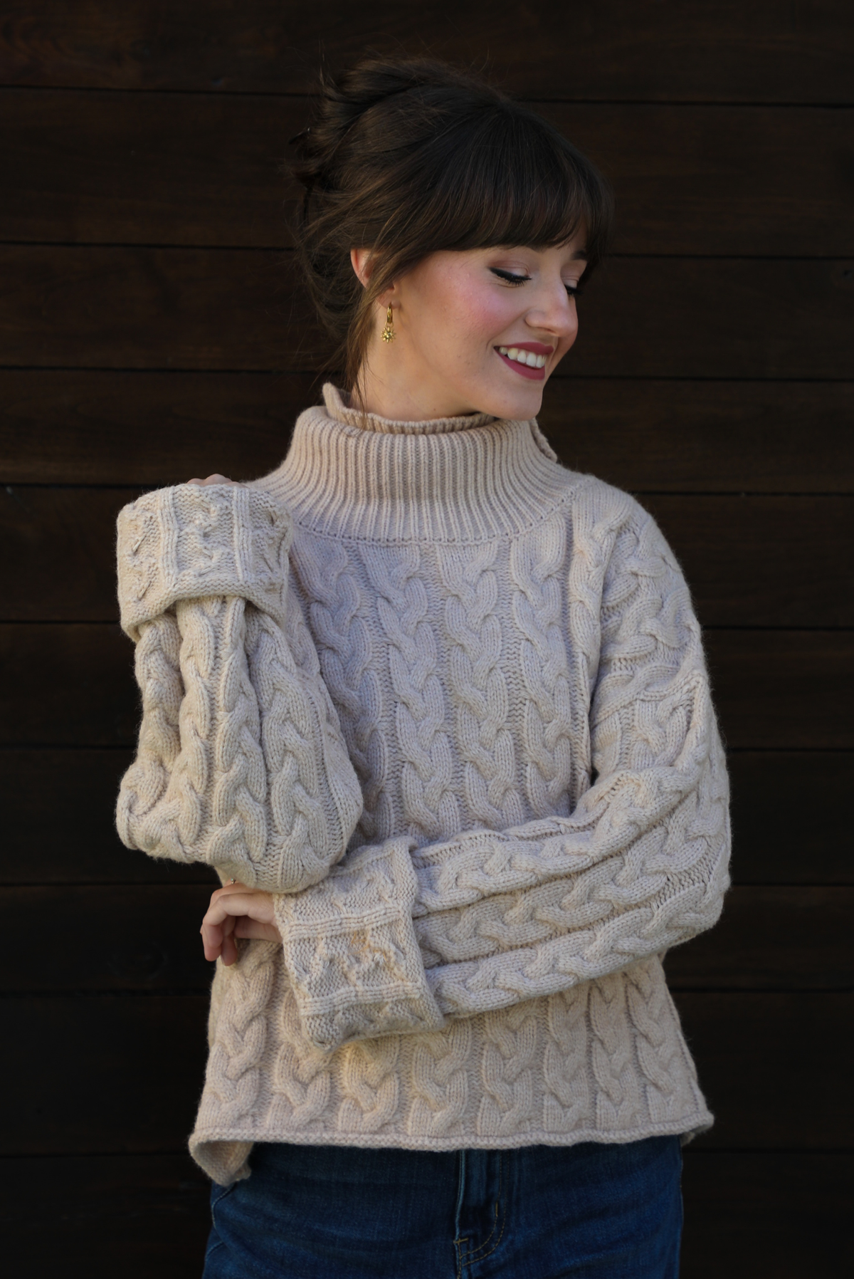 Woman model wearing a tan, thick, cable knit turtle neck sweater with dark denim jeans outdoors in front of a dark stained wooden door. 