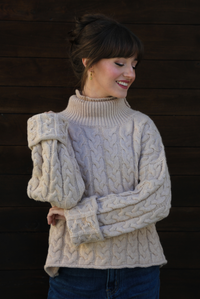 Woman model wearing a tan, thick, cable knit turtle neck sweater with dark denim jeans outdoors in front of a dark stained wooden door. 